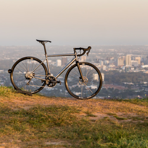 A titanium Bossi gravel/adventure bike on a hill, overlooking the city of Adelaide.