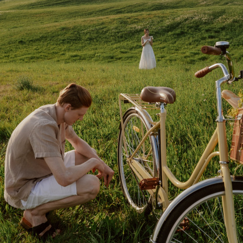 A young man squats down and stares at the chain on his bicycle. He is in a field, a woman in a white dress in the background.