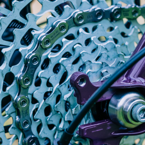 Close-up detail of a Shimano bicycle chain running on a cassette, part of the bicycle's purple frame visible. Copyright Bio-Mechanics Cycles & Repairs.