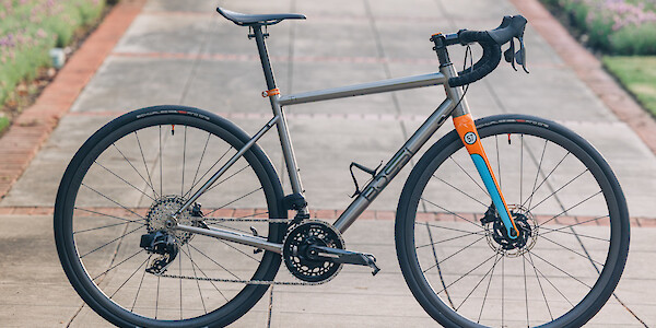 A titanium Bossi Summit endurance bicycle, custom-built by Bio-Mechanics Cycles & Repairs. It's stationed on a brick pathway, flanking flowerbeds in the distance. The fork is painted blue and orange, which stands out against the titanium of the bike.