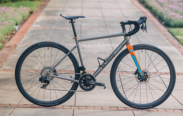 A titanium Bossi Summit endurance bicycle, custom-built by Bio-Mechanics Cycles & Repairs. It's stationed on a brick pathway, flanking flowerbeds in the distance. The fork is painted blue and orange, which stands out against the titanium of the bike.