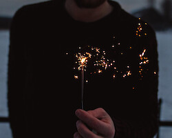 A man holding a sparkler. He is wearing a black jumper, a body of water behind him.