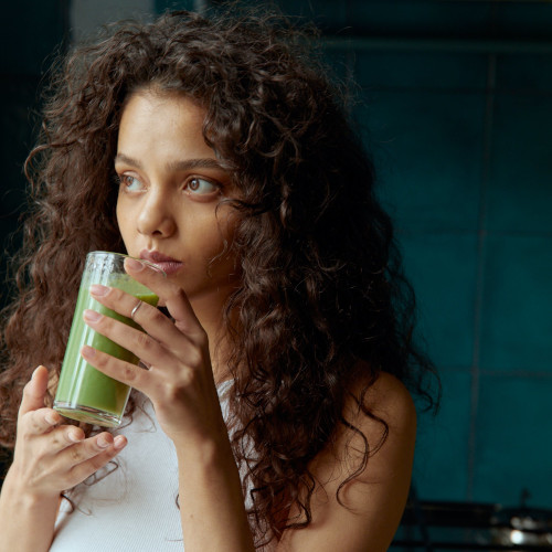 A woman with curly hair, holding a green smoothie up to her lips, looking dubious.