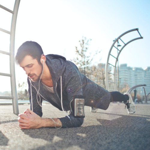 A young man, listening to headphones, holds a plank position on the footpath.
