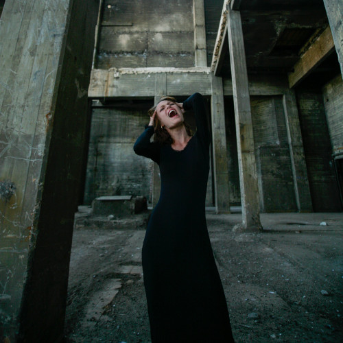 A woman in a black dress, screaming, standing in an abandoned warehouse.