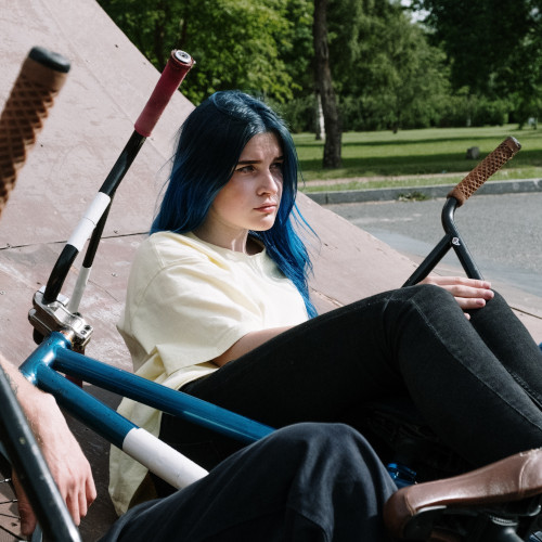 A teenage girl with blue hair sits at a BMX park, looking annoyed.