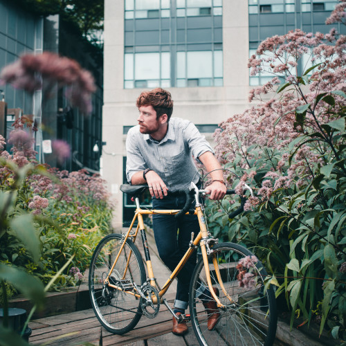 A young man leans against a vintage bicycle in a city garden, surrounded by pink and green flowers.