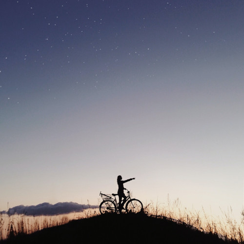 A woman silhouetted against a gentle sunset, standing with her bicycle at the top of a hill, pointing into the distance.