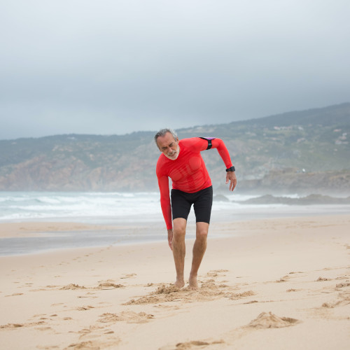 An older man on a beach, bent over in pain and holding his leg.