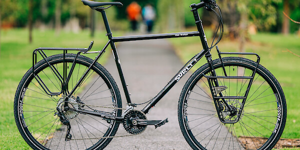 A Surly Disc Trucker steel touring bike, custom-built by Bio-Mechanics Cycles & Repairs. It's standing on a park footpath, people in the distance.