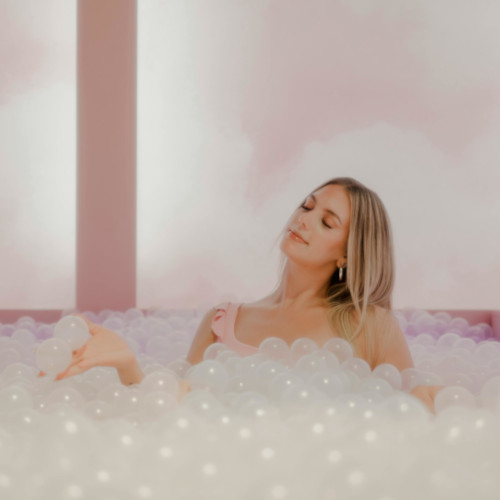 A woman in a pink top, sitting in a bath of transparent balls/bubbles and looking blissful.