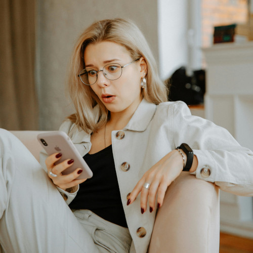 A woman, sitting casually on a couch, looks aghast at something on her phone.