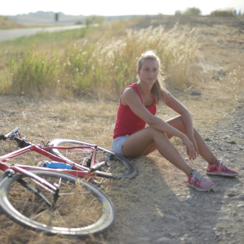 A young woman sitting on the sandy ground next to her bicycle, staring off into the distance.