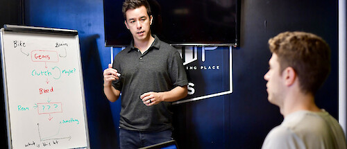 A young man stands next to a whiteboard covered in scribble, explaining a concept to another young man, seated to the right and facing the board.