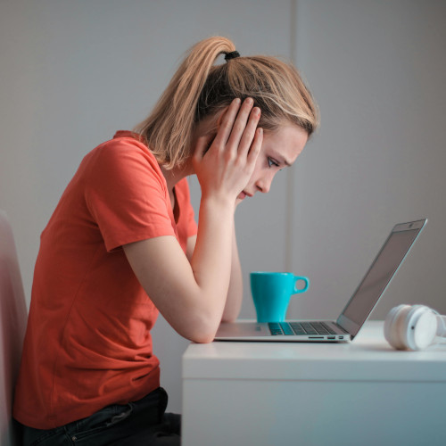 A young woman sits with her head in her hands, staring despondently at a laptop screen.