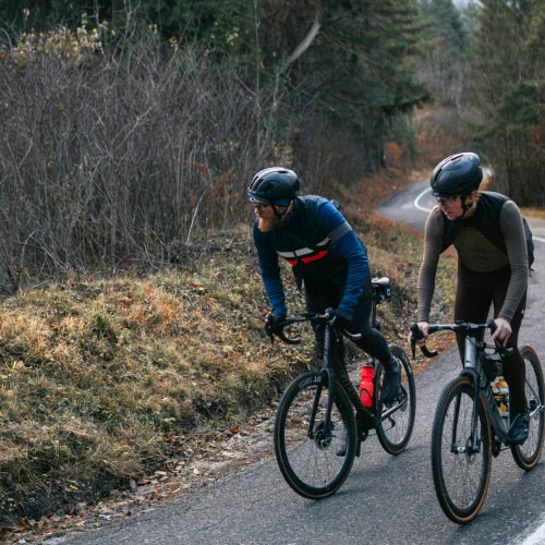 Two cyclists climbing a pathway in a wooded area, both looking off to the side in the same direction.