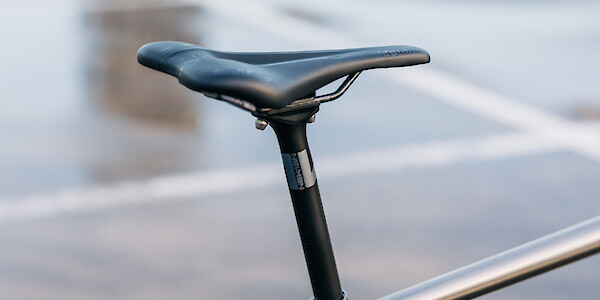 Newmen Advanced carbon bicycle seat post, with a saddle on top, fitted to a titanium Bossi bicycle. Photographed outdoors in a wet car park, visible in the background.