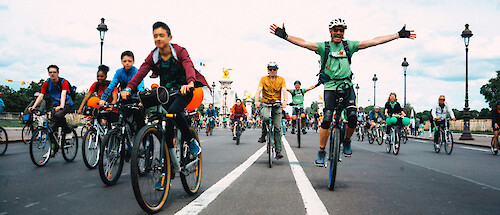 A bunch of people riding bikes along the road towards the camera. One man has his arms spread out in an expression of joy.