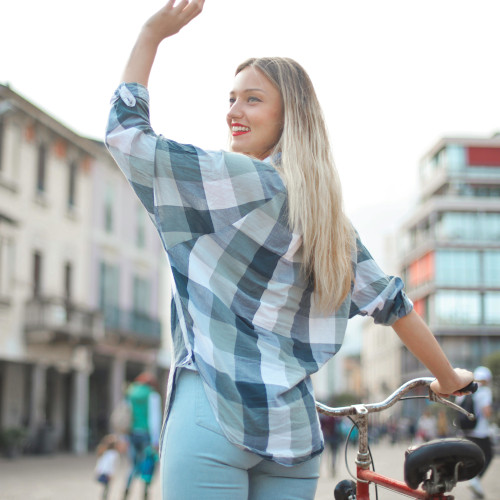 A woman wheeling a bike along a city street, turning back to wave to someone behind her.