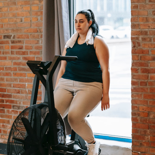 A woman sitting on an exercise bike in the gym, looking unimpressed and tired.