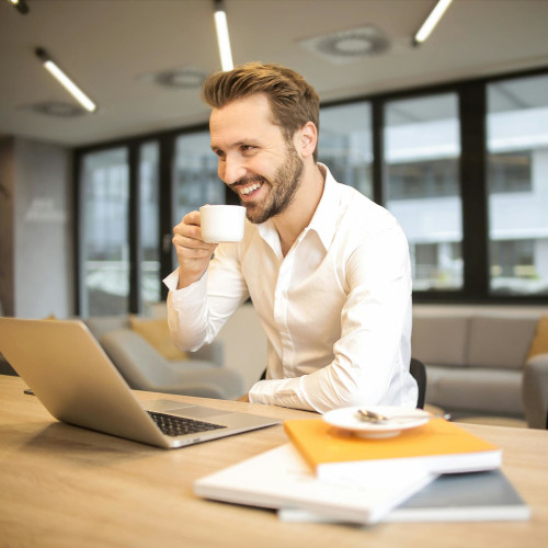 A bearded man sitting at a desk in front of a laptop, drinking a coffee and looking very pleased with himself.