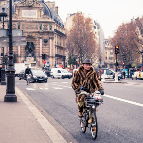 A man riding a bike in a city street. He's wearing a fur coat and a jaunty hat.