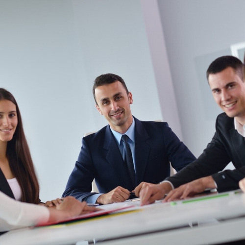 A group of young professionals sitting at a boardroom table, smiling at the screen, looking mildly insincere.