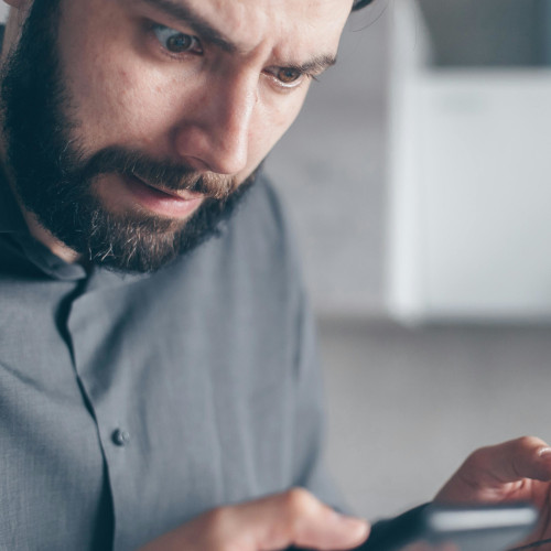 A bearded man looks at his phone screen in wide-eyed horror.