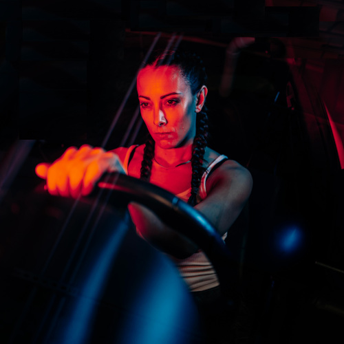 A woman sitting in the driver's seat of a car, looking very cross. She is lit by red neon.