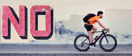 A young man rides a bike along a city street, the word 'NO' graffitied behind him on the wall.