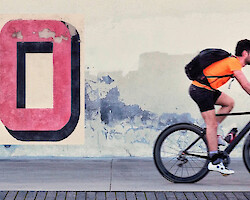 A young man rides a bike along a city street, the word 'NO' graffitied behind him on the wall.