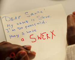 A man writes a letter to Santa. The view is over his shoulder, focused on the letter, which requests a 'SWERX' bike.