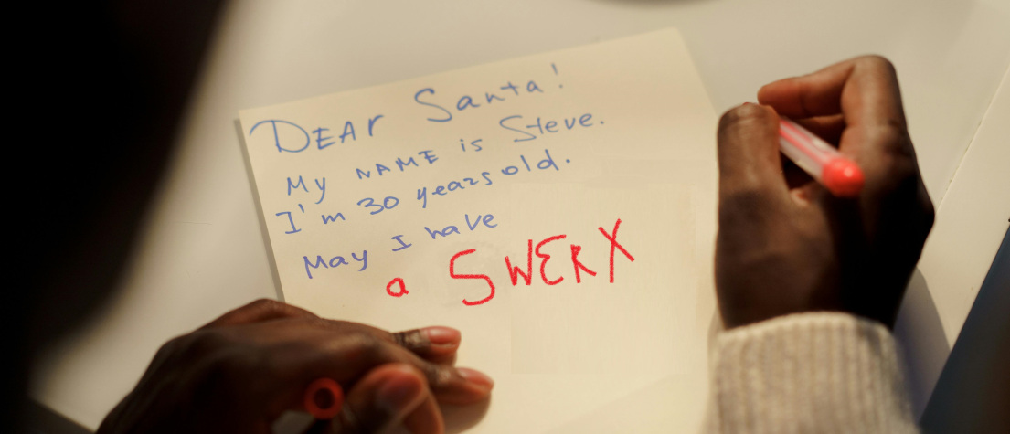 A man writes a letter to Santa. The view is over his shoulder, focused on the letter, which requests a 'SWERX' bike.