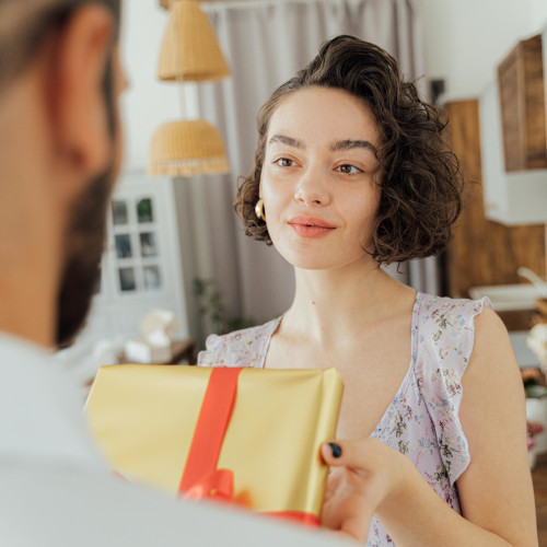 A young woman presents a present wrapped in gold paper to a bearded man.