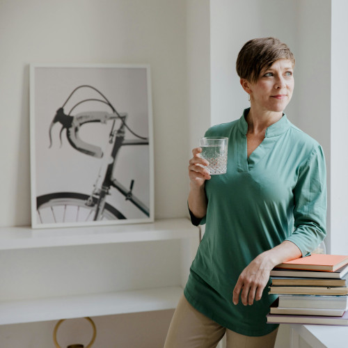 A woman drinking a glass of water, leaning on a stack of books and looking out a window. A bicycle print is in view behind her.