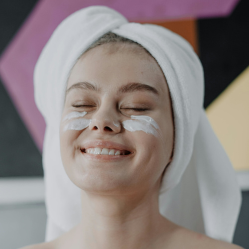 A smiling young woman with moisturiser on her cheeks, her hair in a towel.
