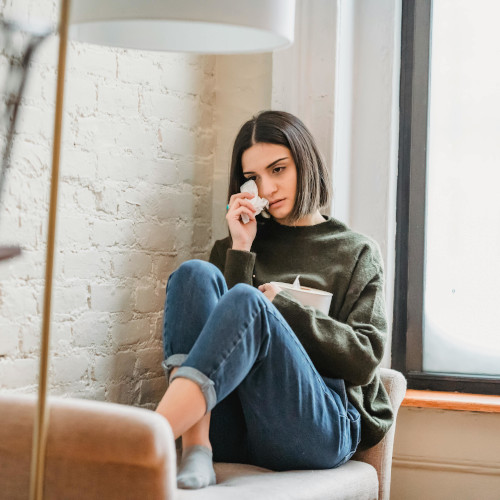 A girl sits by a window seat, dabbing away tears with a tissue.