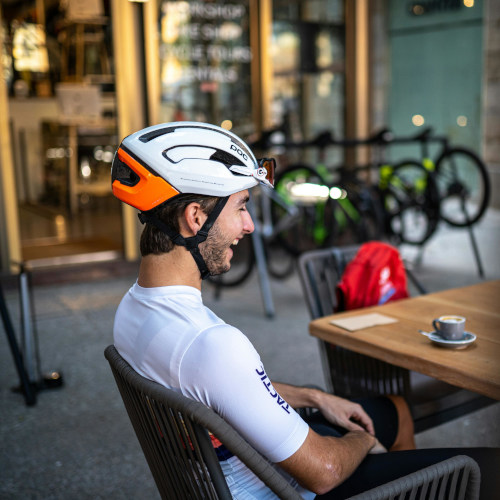 A young man in cycling kit sitting at a cafe, laughing. There's a coffee in front of him.