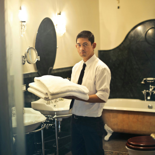 A man in a shirt and tie, holding a stack of towels in a fancy bathroom, looking slightly annoyed.