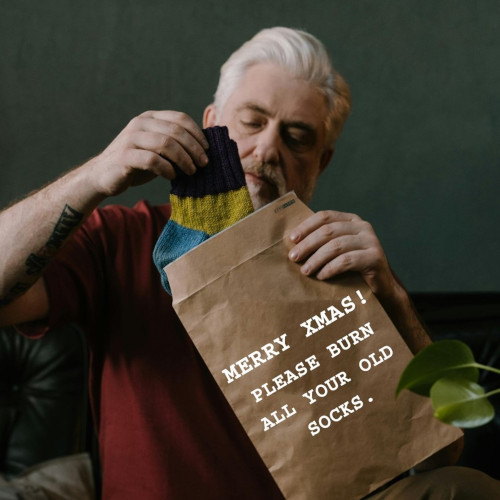 An old man pulls a pair of colourful woolen socks out of a paper bag. The bag reads 'Merry Xmas! Please burn all of your old socks.'