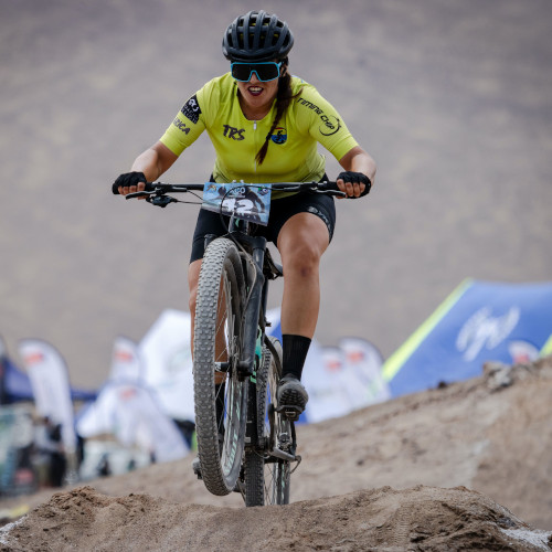 A woman competiting in a mountain bike race, mid-air over a jump.