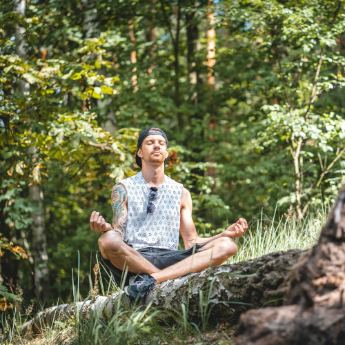 A young man sitting cross-legged on a log in the wilderness, meditating. He is wearing a backwards baseball cap.