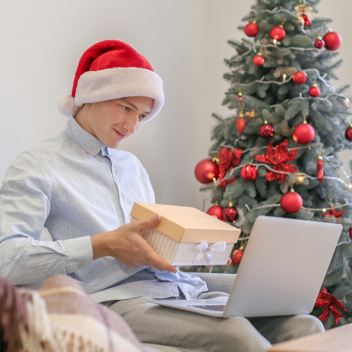 A young man wearing a Santa hat, sitting next to a tree and offering a wrapped gift to the laptop on his knees.