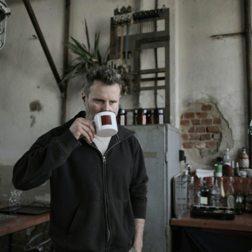 A bearded man standing in a kitchen, drinking from a mug and looking like he's just realised something.