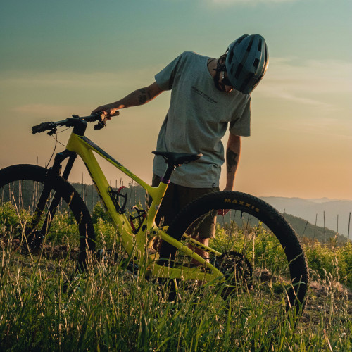 A young man standing in a field next to his mountain bike, looking at the back tyre in dismay.