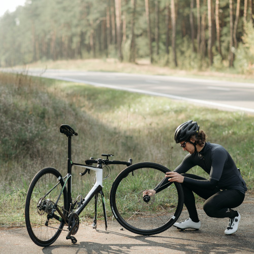 A young man in lycra cycling gear, crouched down to work on his bicycle wheel by the side of the road.