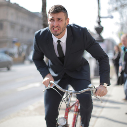 A young man in a suit, riding an old bicycle along a city street, wearing an expression of pain.