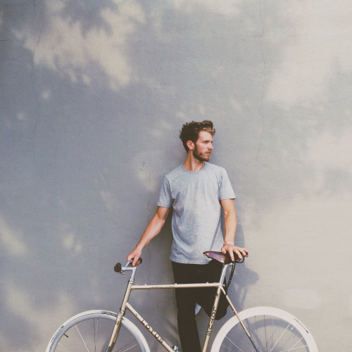 A young man leaning against a pale grey wall, looking into the distance and holding a bicycle in front of him.