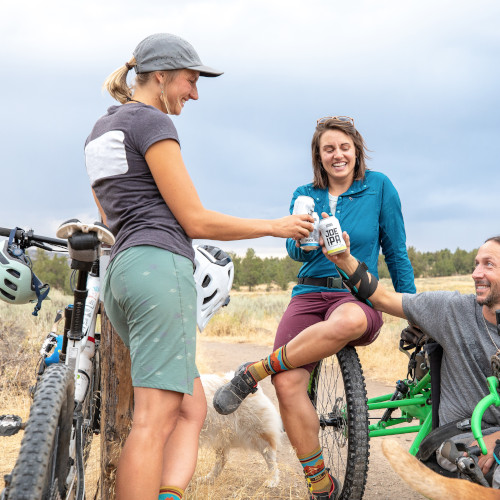 Three mountain bikers (two women, one man) toasting each other with beer cans after a ride, smiling.