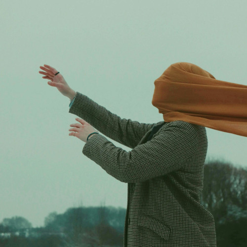 A person with a scarf over their face, facing into a high wind, arms reaching out in front of them.